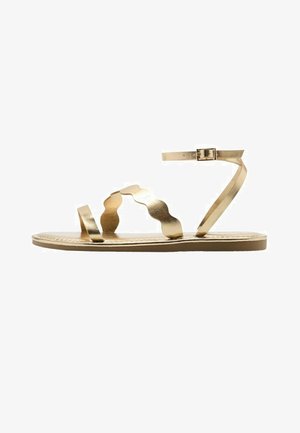 Platte metalen gouden sandaal met enkelbandje en golvende middenvoetband op een dunne zool, zijaanzicht op witte achtergrond.