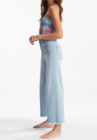 Jeans de pierna ancha azul claro con dobladillo deshilachado, combinados con una blusa sin mangas de patrones multicolores. La tela parece tener una textura y ser elástica.