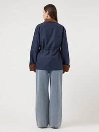 Γυναίκα που στέκεται με την πλάτη στραμμένη, φορώντας ένα ναυτικό σακάκι με καφέ γιακά και μανσέτες, σε συνδυασμό με φαρδιά ανοιχτό μπλε τζιν, σε μια απλή φόντο.