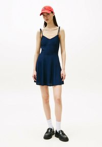 Vestido mini azul marino con tirantes finos, cuerpo ajustado y falda acampanada. La modelo lleva una gorra roja y mocasines negros con calcetines blancos.