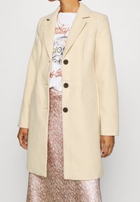 Cappotto in misto lana beige con rever a punta, design monopetto e quattro bottoni scuri. Indossato sopra una T-shirt grafica e una gonna floreale.