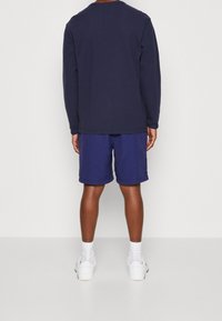 Námořnické tričko s dlouhým rukávem v kombinaci s modrými šortkami; oboje vyrobené z měkké látky. Pro sportovní vzhled jsou obuty bílé tenisky a ponožky.