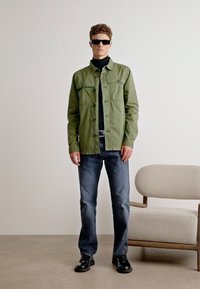 Olivově zelená lehká bunda se dvěma předními kapsami, černý rolák, modré džínové kalhoty a černé boty. Model stojí na jednotném pozadí.