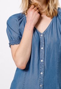 Blusa de mezclilla en azul claro con mangas cortas abullonadas y escote en V. Cuenta con cierre delantero de botones con botones blancos a juego y detalle de mangas fruncidas.