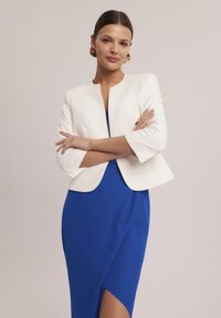 Biała krótka marynarka nałożona na niebieską sukienkę z rozcięciem do uda. Marynarka ma okrągły dekolt i rękawy do trzech czwartych. Złote kolczyki podkreślają stylizację.