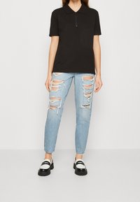 Kvinna som bär en svart kortärmad pikétröja, ljusblå ripped jeans och svart-vita chunky loafers som står framför en vit bakgrund.