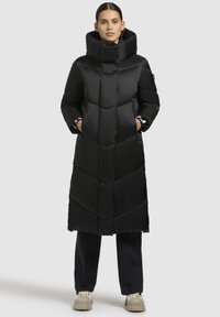 Svart puffjacka med ett sicksackmönster, hög krage och sidofickor. Slätt, glänsande material. Kombinerad med lösa svarta byxor och beige skor.