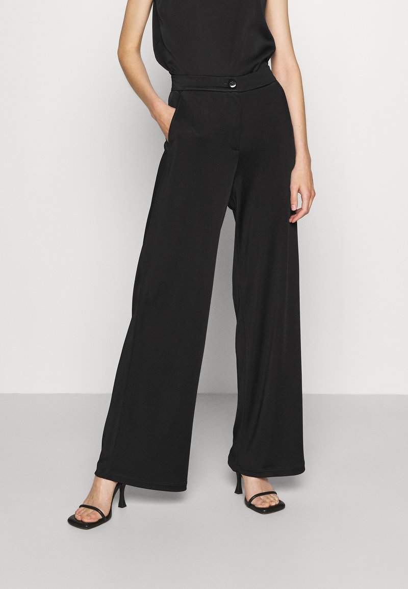 Model draagt een zwarte mouwloze top, zwart wijde broek met hoge taille en knoopdetails, en zwarte sandaalhakken met bandjes, staand met hand in de zak.