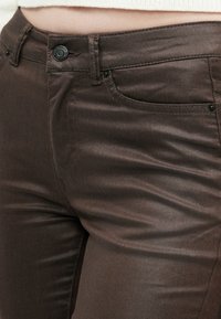 Detaily hnedých tesne priliehavých nohavíc ukazujúce predné vrecko, pútka na opasok a gombíkový uzáver v páse na holých nohách.