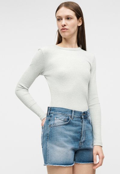 Νεαρή γυναίκα με μακριά καστανά μαλλιά, που φοράει γκρι ανοιχτό μακρυμάνικη ριμπ μπλούζα και μπλε τζιν σορτς, μπροστά σε απλό φόντο.