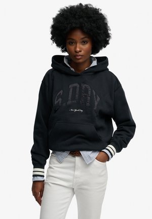 Kobieta o kręconych włosach, ubrana w czarną bluzę z kapturem S.DRY na niebieską koszulę w paski i białe spodnie, stojąca z jedną ręką w kieszeni na białym tle.