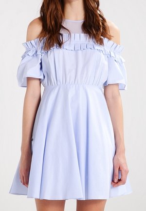 Femme portant une robe bleu clair à épaules dénudées avec un col à volants et une taille cintrée, debout devant un fond uni.