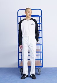 Camicia a maniche lunghe bianca e nera con un logo, pantaloni bianchi e sneakers nere con strisce bianche. Lo sfondo presenta un carrello blu.