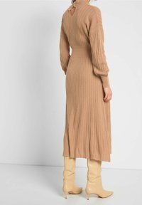 Stickad klänning i beige med ribbad textur; har långa ärmar, en åtsittande midja och en nyckelhålsöppning i ryggen. Matchas med knähöga beige stövlar.