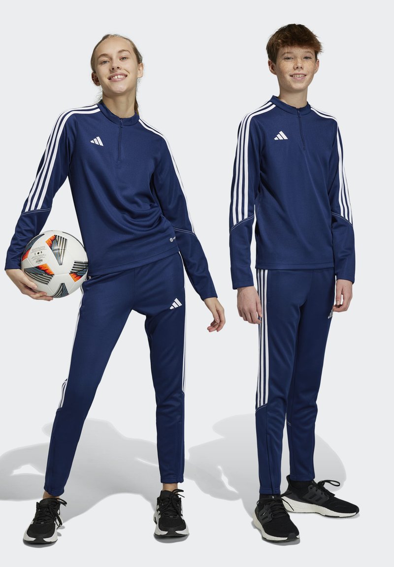 Marineblaue Trainingsanzüge mit weißen Streifen, featuring lange Ärmel und Reißverschluss am Kragen. Ein Model hält einen Fußball. Schwarze Turnschuhe.