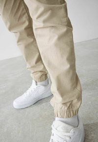 Beige, kevyet housut, joissa on elastiset hihnat ja sileä pinta, yhdistettynä valkoisiin, matalavartisiin Nike-kengiin, joissa on rei'ityksiä.