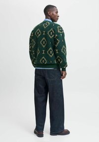 Pull en maille verte à motifs de diamants, associé à un pantalon en denim large bleu foncé. Le pull a une finition texturée et des poignets côtelés.