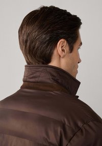 Chaqueta acolchada marrón con cuello alto, que presenta una textura suave y una marca discreta cerca del cuello. Detalles de costuras visibles.