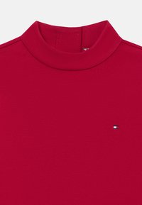 Haut rouge à col montant fabriqué dans un matériau lisse et extensible. Présente un petit logo avec des accents bleus, blancs et rouges sur la poitrine.