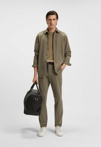 Olijfgroene blouse en broek van textuurstof, gecombineerd met een beige top en witte sneakers. Zwarte duffeltas in één hand vastgehouden.