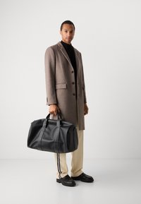 Bolso de viaje de cuero negro con dos asas y costuras en contraste. Textura suave, gran capacidad y forma redondeada que ofrecen funcionalidad.