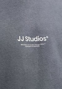 Γκρι ύφασμα με λευκό κείμενο λογότυπου "JJ Studios," και μικρότερο κείμενο "BROUGHT TO YOU BY THE J&J CREW, DESIGNED IN DENMARK" από κάτω.