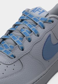 Baskets Nike Air Force 1 en cuir gris avec des lacets bleus portant la marque Nike et un logo swoosh Nike bleu sur le côté.