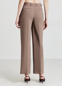 Pantalon pattes d'éléphant marron clair avec une texture lisse. Comprend deux poches arrière et une taille mi-haute. Associé à des talons rouges foncés.