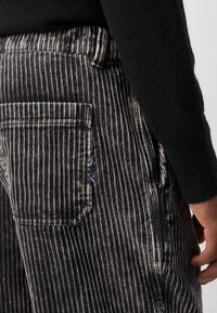 Gestreepte corduroy broek in zwart en grijs met een textuurafwerking. Heeft een achterzak en een blauw borduurdetail op de tailleband.