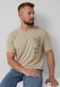 Beige katoenen t-shirt met een boomgrafiek in donkerbruin, ronde halslijn en korte mouwen. Gecombineerd met lichtblauwe spijkerbroek.