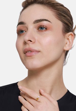 Goldring mit geometrischem Design und einem kleinen Perlelement. Das Modell trägt ein schwarzes Oberteil, das ihre glatte Haut und das dezente Makeup zur Geltung bringt.