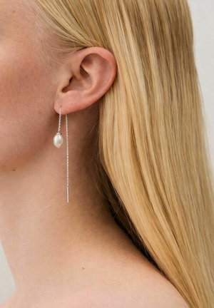 Femeie blondă purtând o cercei cu lanț argintiu și o perlă ovală albă agățată la urechea stângă, pe un fundal neutru.