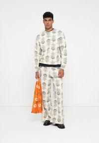 Hombre vistiendo un conjunto de color crema a juego con un patrón floral negro, sosteniendo una bolsa de tote floral naranja, de pie frente a un fondo blanco.