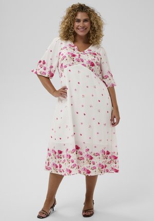 Femme debout souriante, portant une robe midi blanche avec des motifs floraux roses et des talons ouverts noirs, sur un fond clair uni.