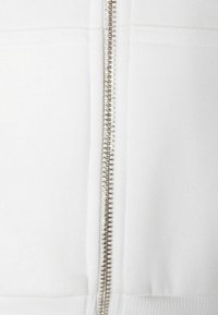 Primo piano di una cerniera in metallo argentato su un indumento in tessuto bianco con cuciture visibili e orlo a coste sul fondo.