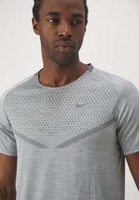 Hallitusäiteline hall sportlik t-särk tekstuurse mustriga, ümmarguse kaelusega ja halli Nike logo kaunistusega. Kujunduses on kontrastsed chevroni aktsendid.