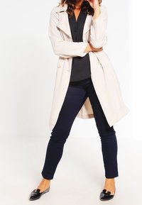 Lichtbeige trenchcoat met grote revers en knoopaccenten, gecombineerd met navy fitted broek en glanzende zwarte ballerina's, tegen een witte achtergrond.