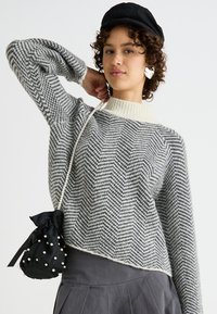 Sweater s uzorkom riblje kosti u sivoj i bijeloj boji s kremastim rebrastim ovratnikom, nosi se uz plisiranu sivu suknju i crnu torbicu s teksturom koja ima bisere.