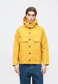 Gul hettejakke laget av teksturert stoff, med brune knapper, to frontlommer og en avslappet passform. Brukt med blå denim bukser.