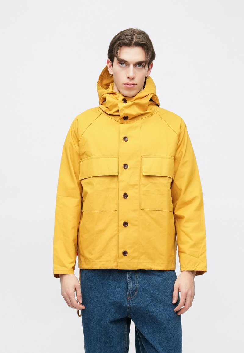 Gul hettejakke laget av teksturert stoff, med brune knapper, to frontlommer og en avslappet passform. Brukt med blå denim bukser.