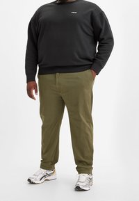 Schwarzer Rundhals-Sweatshirt mit weißem Logo, olivgrüne Chinos und silberne Sportschuhe mit schwarz-goldenen Akzenten.