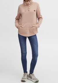 Sweat à capuche rose clair avec un motif à pois, poche avant et cordon de serrage. Porté avec un jean skinny bleu foncé et des baskets beige.