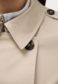 Beżowy trencz z błyszczącym brązowym guzikiem i usztywnianym kołnierzem, wyposażony w zamek błyskawiczny oraz miękką, jasnoszarą podszewkę.