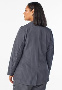 Blazer gris a rayas con mangas largas, cuello de muesca y una apertura trasera. Presenta un ajuste entallado y una textura suave.