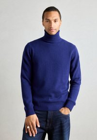 Herren Rollkragenpullover in einem satten Marineblau, aus weichem Material mit gerippten Bündchen und Saum. Getragen über dunklen Denim-Jeans.