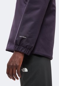 Main à la peau foncée portant une veste violet foncé avec "DRYVENT" sur la manche et un pantalon noir avec le logo "The North Face" sur la cuisse.