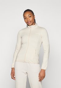 Beige taillierte Jacke mit hohem Kragen, Reißverschluss vorne und gerippten Seitenpaneelen; weicher, strukturierten Stoff mit zwei Reißverschlusstaschen.