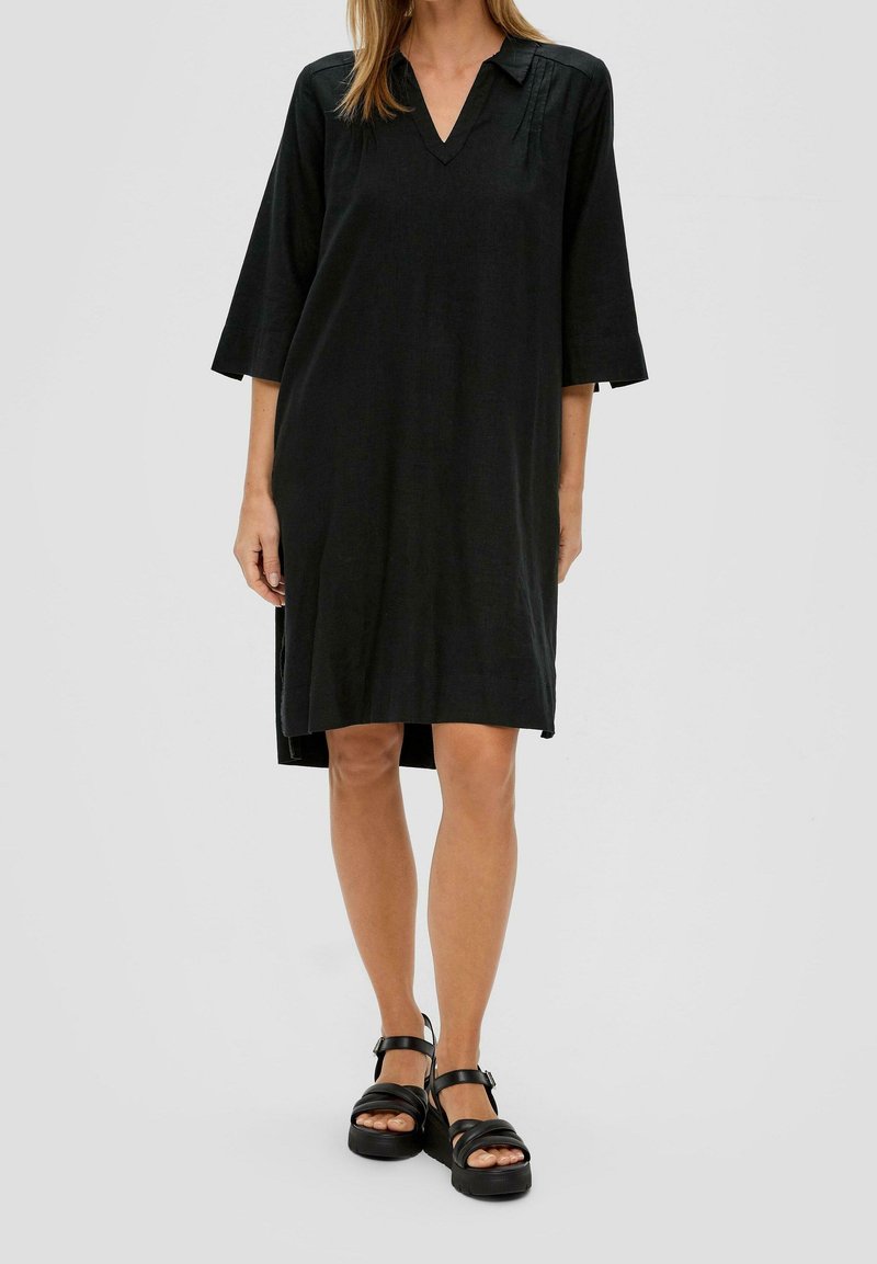Femme portant une robe noire ample longueur genoux avec des manches trois-quarts et des sandales plates-formes noires, debout devant un fond uni.
