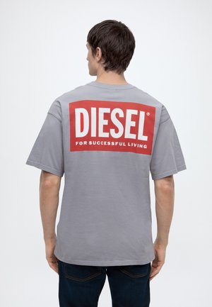 Uomo di spalle che indossa una t-shirt grigia Diesel con un grande logo rosso e bianco stampato sulla schiena e jeans blu scuro.