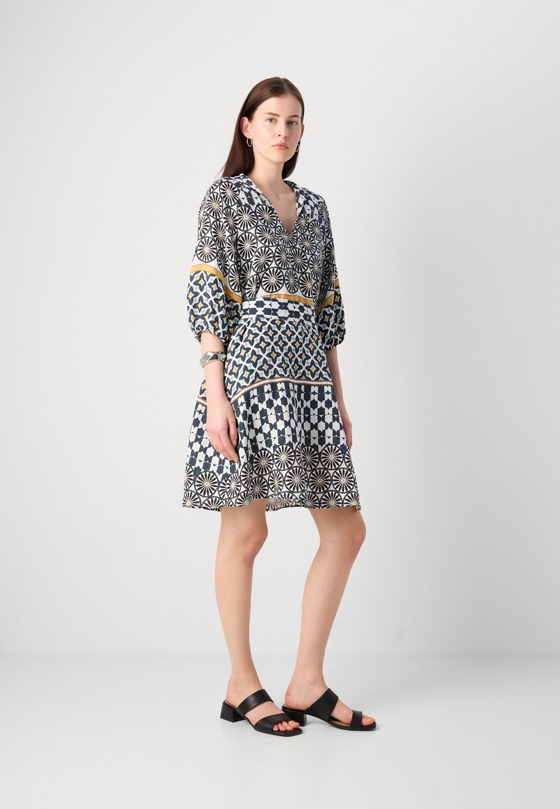 Gemustertes Kleid mit V-Ausschnitt, drei Viertelärmeln und tailliertem Bund. Es hat geometrische Drucke in Marineblau, Weiß und Gelb. Schwarze Sandalen.